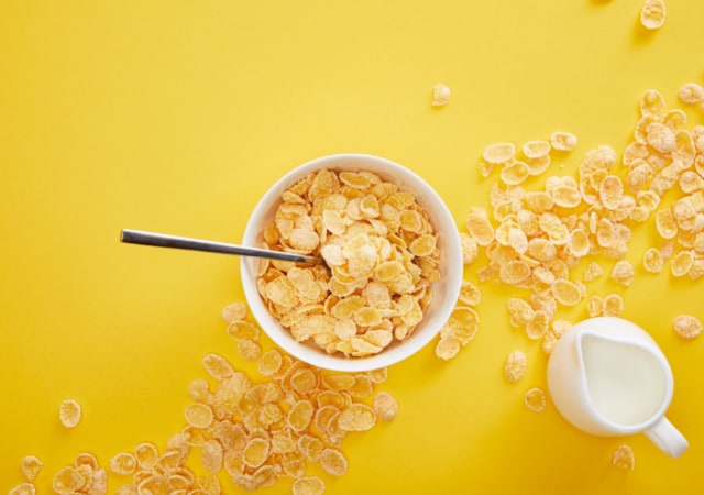 Image of cereal in a bowl and a spoon with milk and cereal grains on a yellow table top