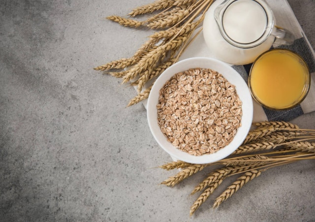 Whole grain cereal in a white bowl with milk, orange juice, and wheat on a table cloth