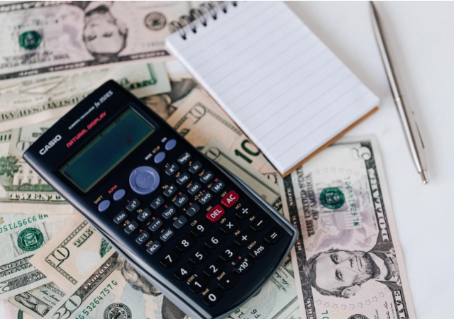 A calculator, pen, and a notepad on top of scattered dollar bills on a table